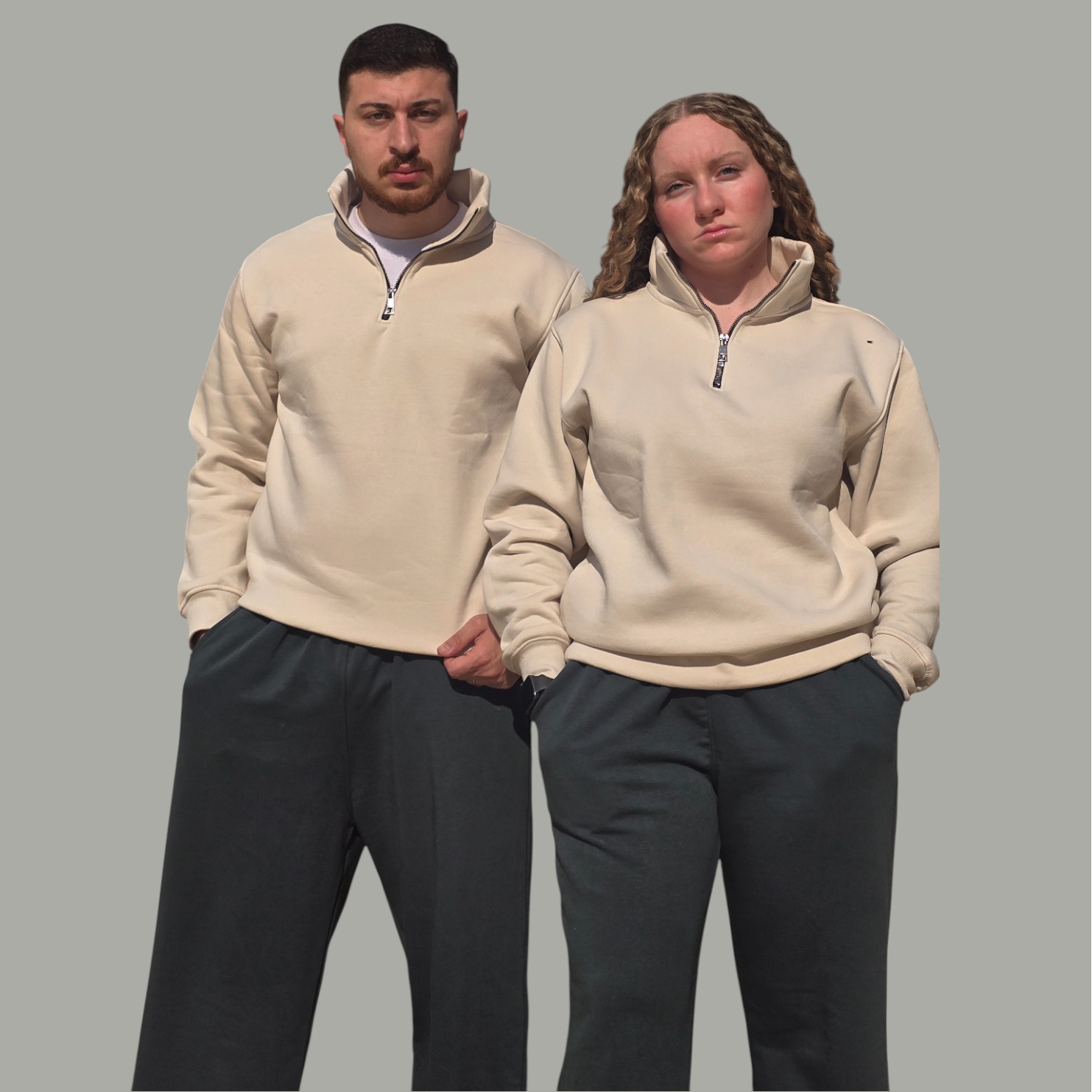 Two people wearing matching beige pullovers and dark pants on a gray background