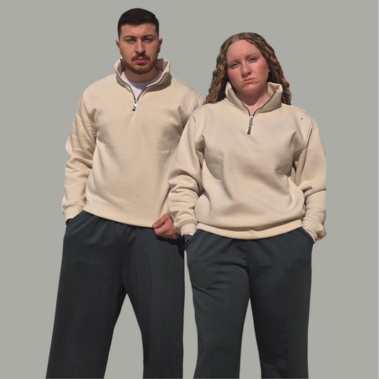 Two people wearing matching beige pullovers and dark pants on a gray background